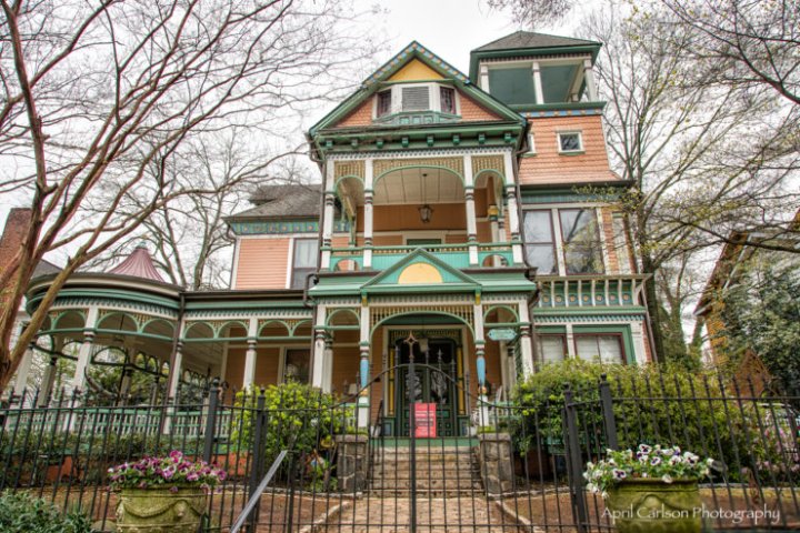Victorian-style house with ornate green and orange trim, surrounded by trees and a black metal fence.