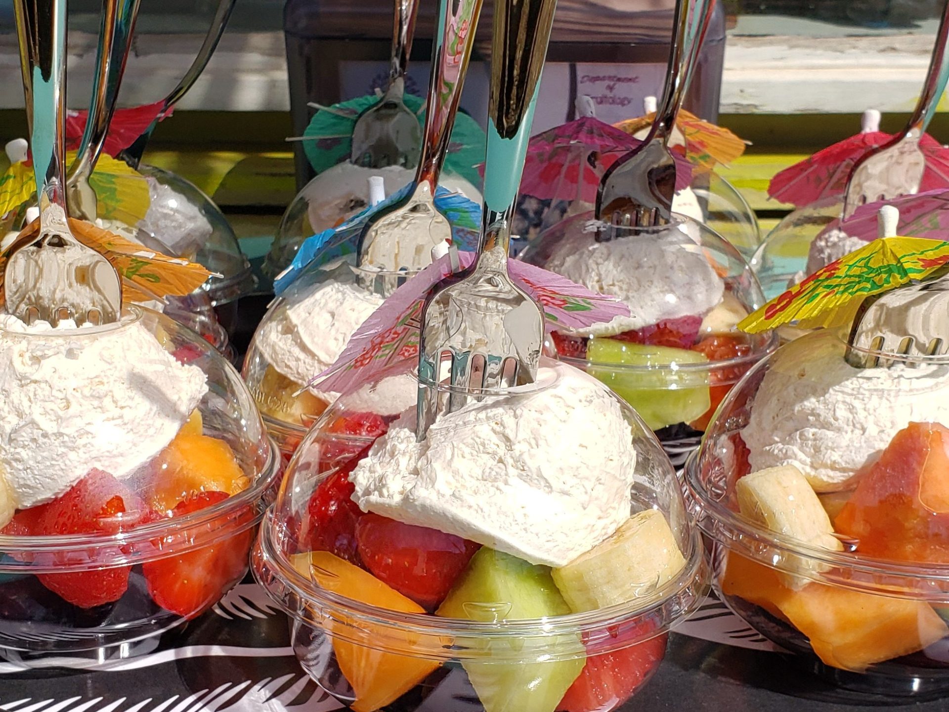 Fruit cups with whipped cream, plastic forks, and umbrellas on a blue table.