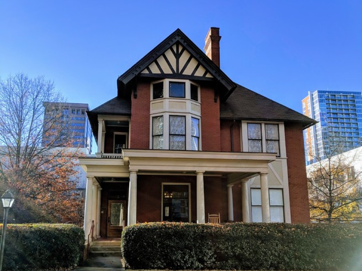 Large brick house with gabled roof and porch, surrounded by city buildings.