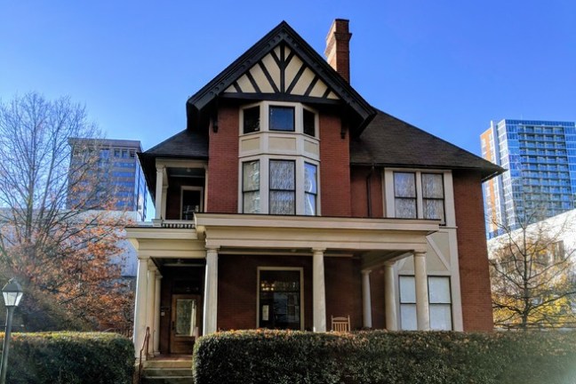 Large brick house with gabled roof and porch, surrounded by city buildings.
