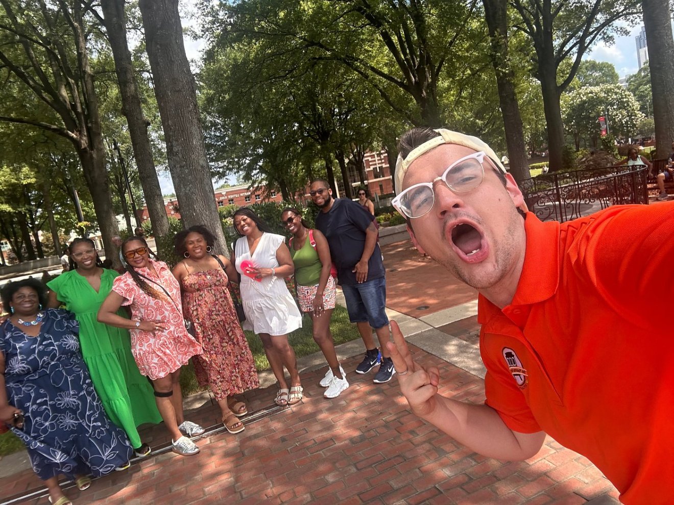 Group selfie of 8 people outdoors on a sunny day with trees in the background.