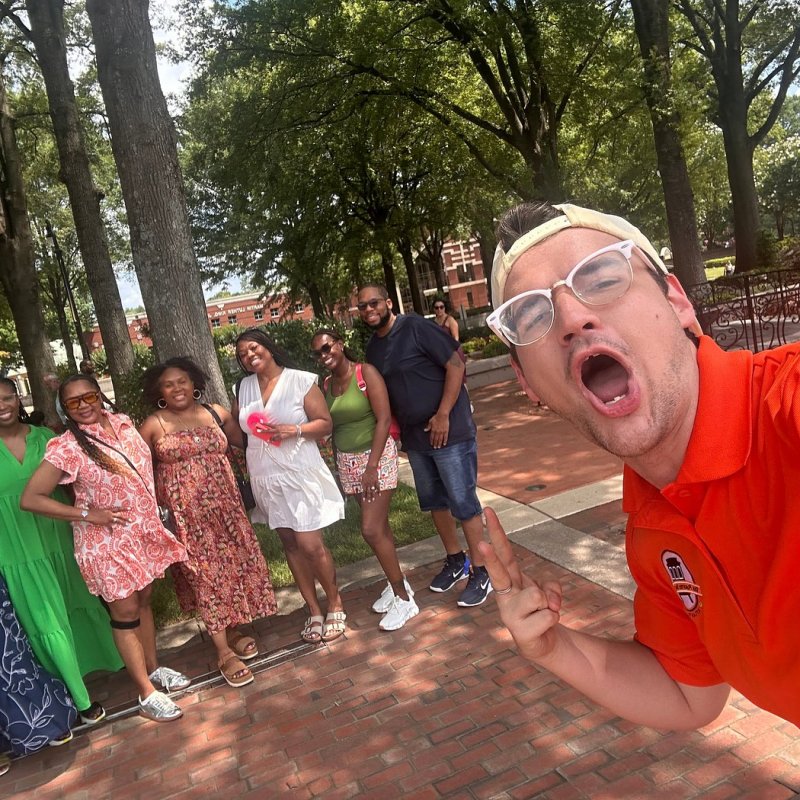 Group selfie of 8 people outdoors on a sunny day with trees in the background.