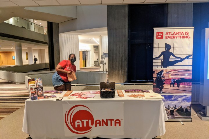 Person in red shirt standing behind a table with Atlanta banner in a lobby, holding up literature.