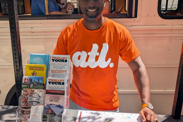 Smiling man in orange 'atl.' shirt beside brochures and maps near a trolley.