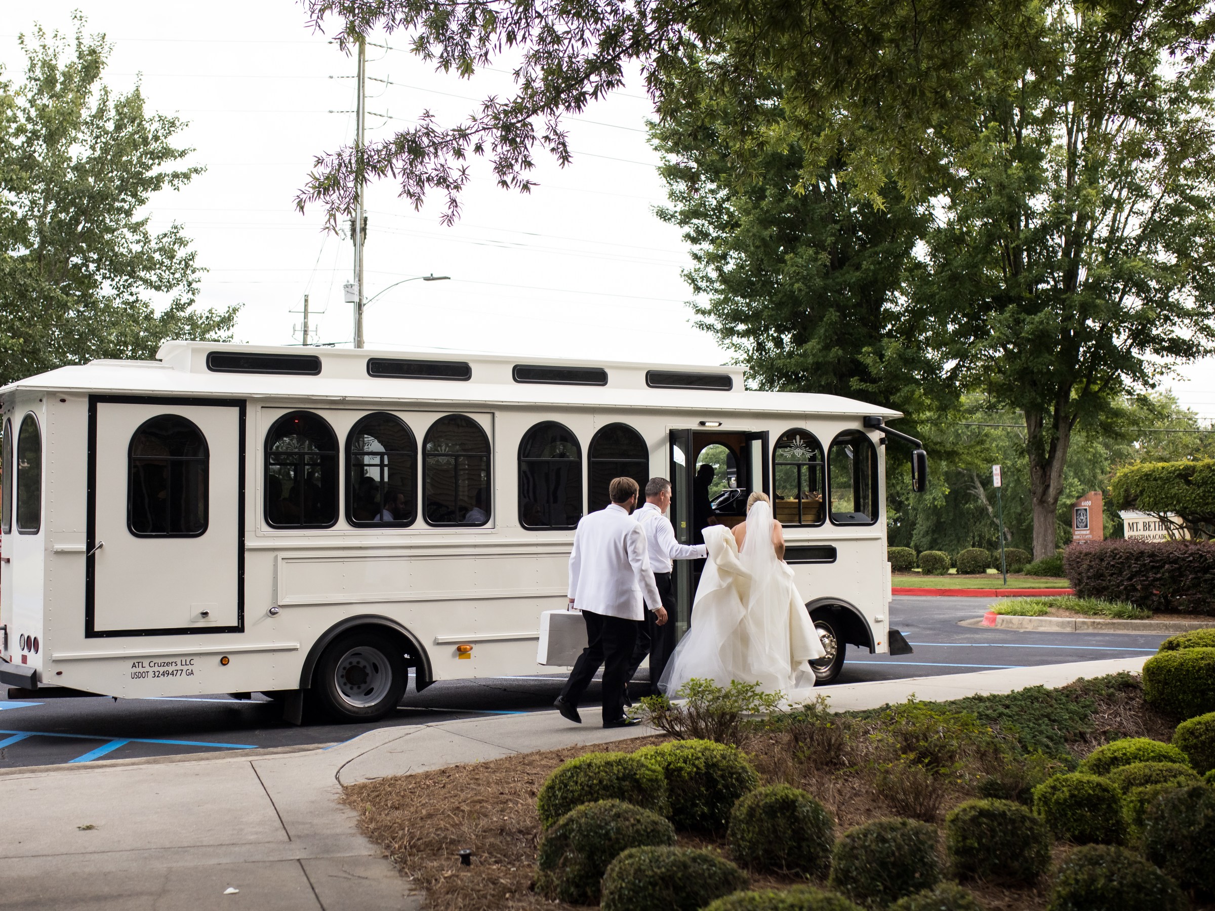 Bride and groom board a white wedding trolley on a tree-lined street.