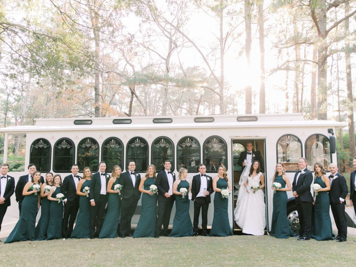 Wedding party in formal attire posing beside a white trolley in a wooded area.