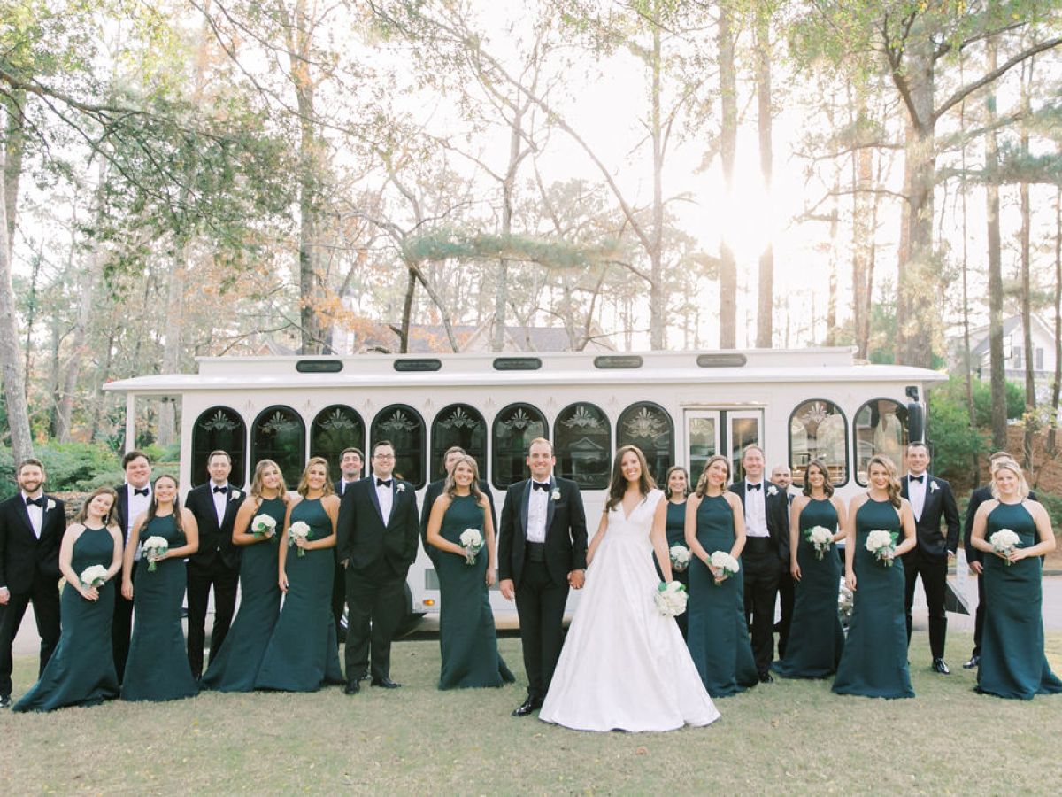 Wedding party poses in front of a white trolley in a wooded area.