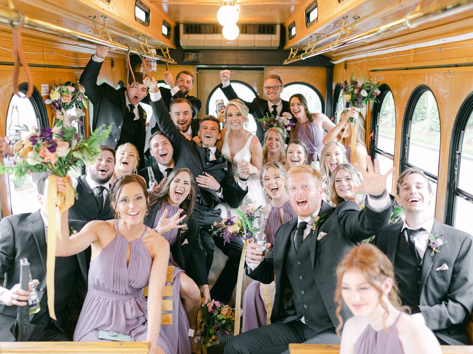 Joyful wedding party posing inside a decorated trolley, with people smiling and holding bouquets.