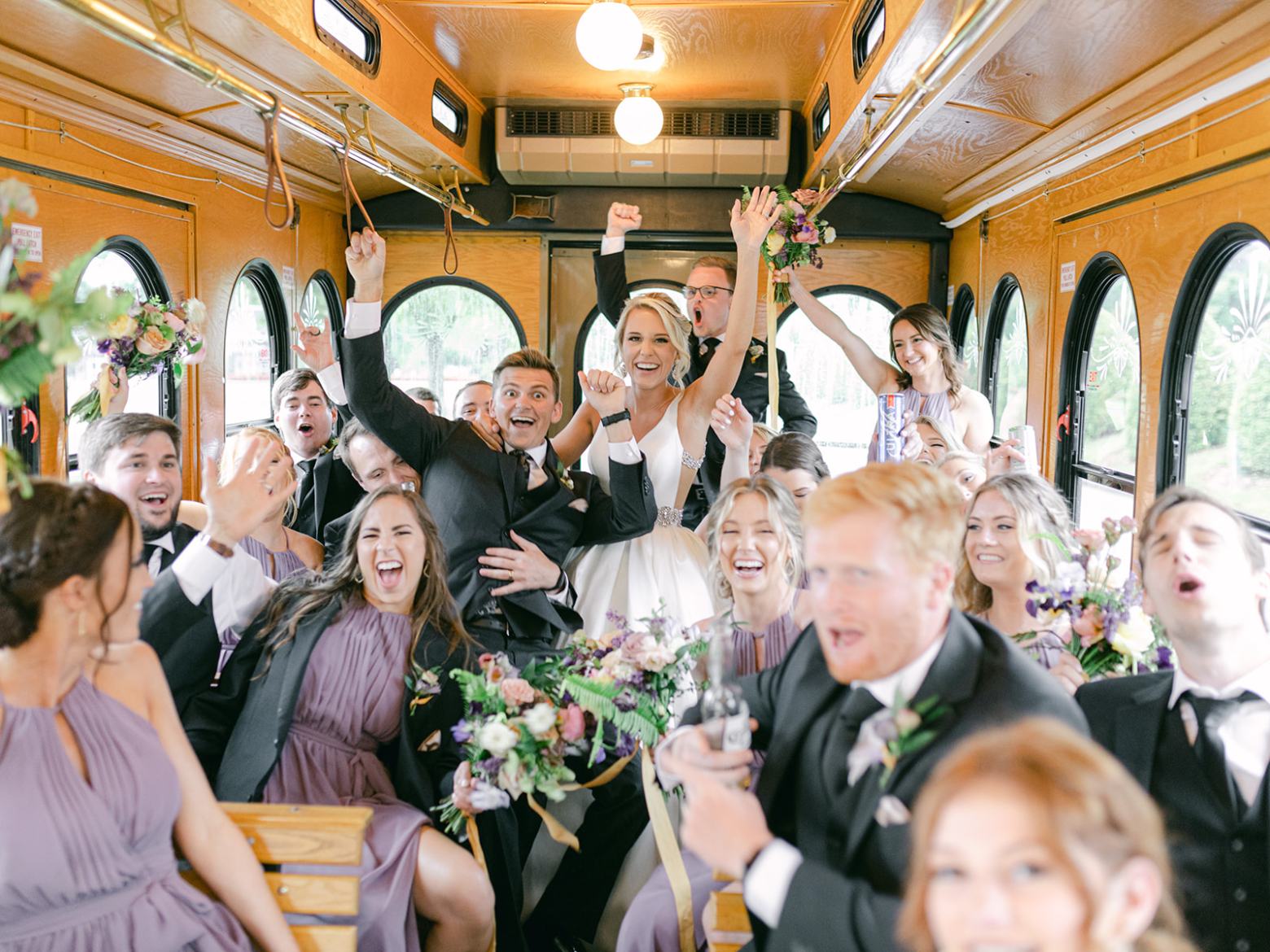 Group of people celebrating on a bus wearing formal attire, with raised arms and joyful expressions.