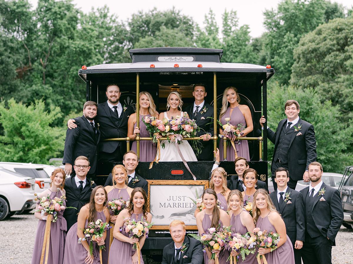 Wedding party in formal attire poses around a 'Just Married' trolley, surrounded by trees.