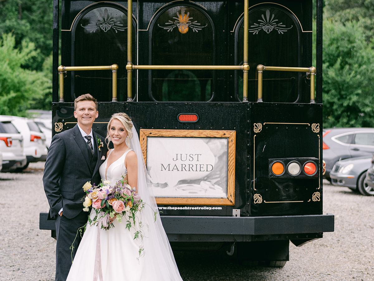 Bride and groom stand in front of a 'Just Married' trolley on a gravel driveway.