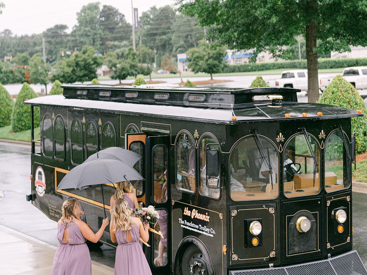 Three women in purple dresses board a black trolley on a rainy day, one holding an umbrella.